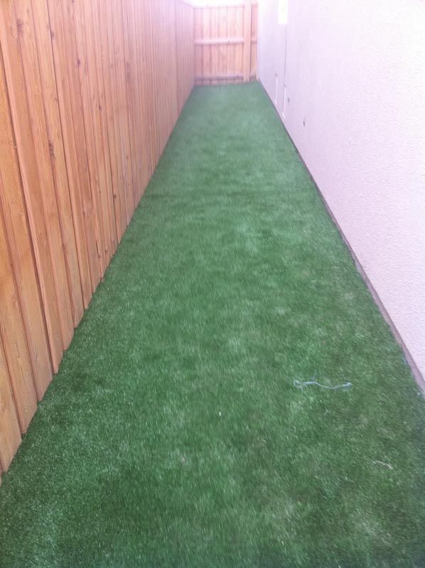 Narrow outdoor walkway with green artificial turf between a wooden fence and a white wall