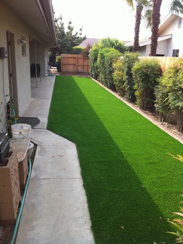 Narrow backyard with bright green artificial turf, concrete walkway, and trimmed hedges beside a house