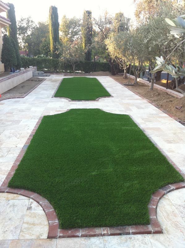 Backyard with two green artificial turf sections bordered by light stone and brick edging, lined with trees