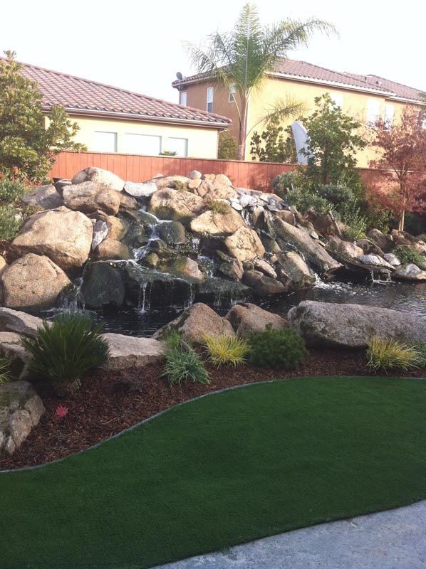 Rock waterfall and boulders in a landscaped front yard with green lawn and palm trees.