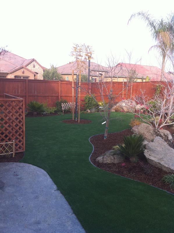 Backyard with green lawn, stone path, wooden fence, and landscaped plants and rocks.