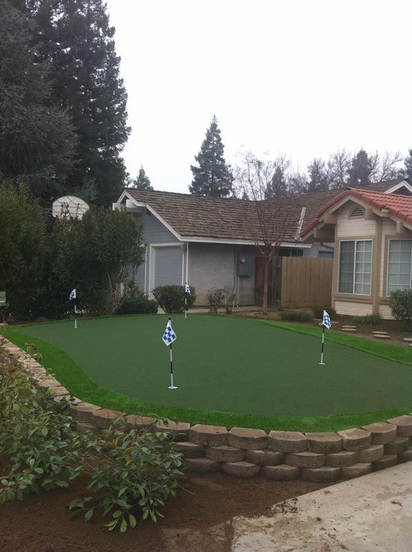 Small backyard putting green with two golf holes beside a house and low brick retaining wall