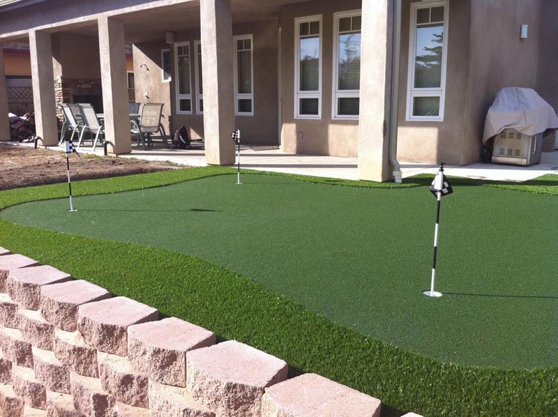 Backyard putting green beside a patio, with a golf flag and house exterior in the background