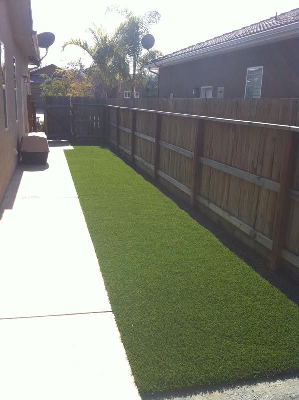 Narrow side yard with artificial grass, concrete path, and wooden fence beside a house.