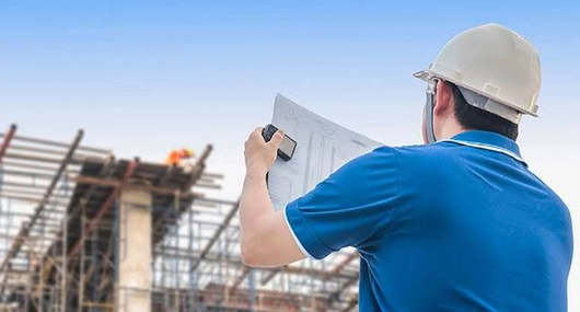 Construction worker in blue shirt and hard hat reviewing plans at a building site.
