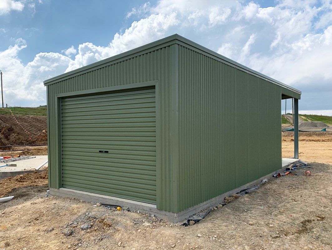 Green metal shed with a roller door on a concrete base, set on a construction site.