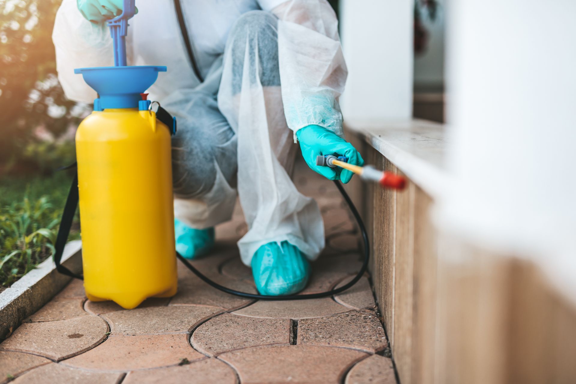 Pest control worker spraying insecticide near a house, wearing protective gear; yellow tank.