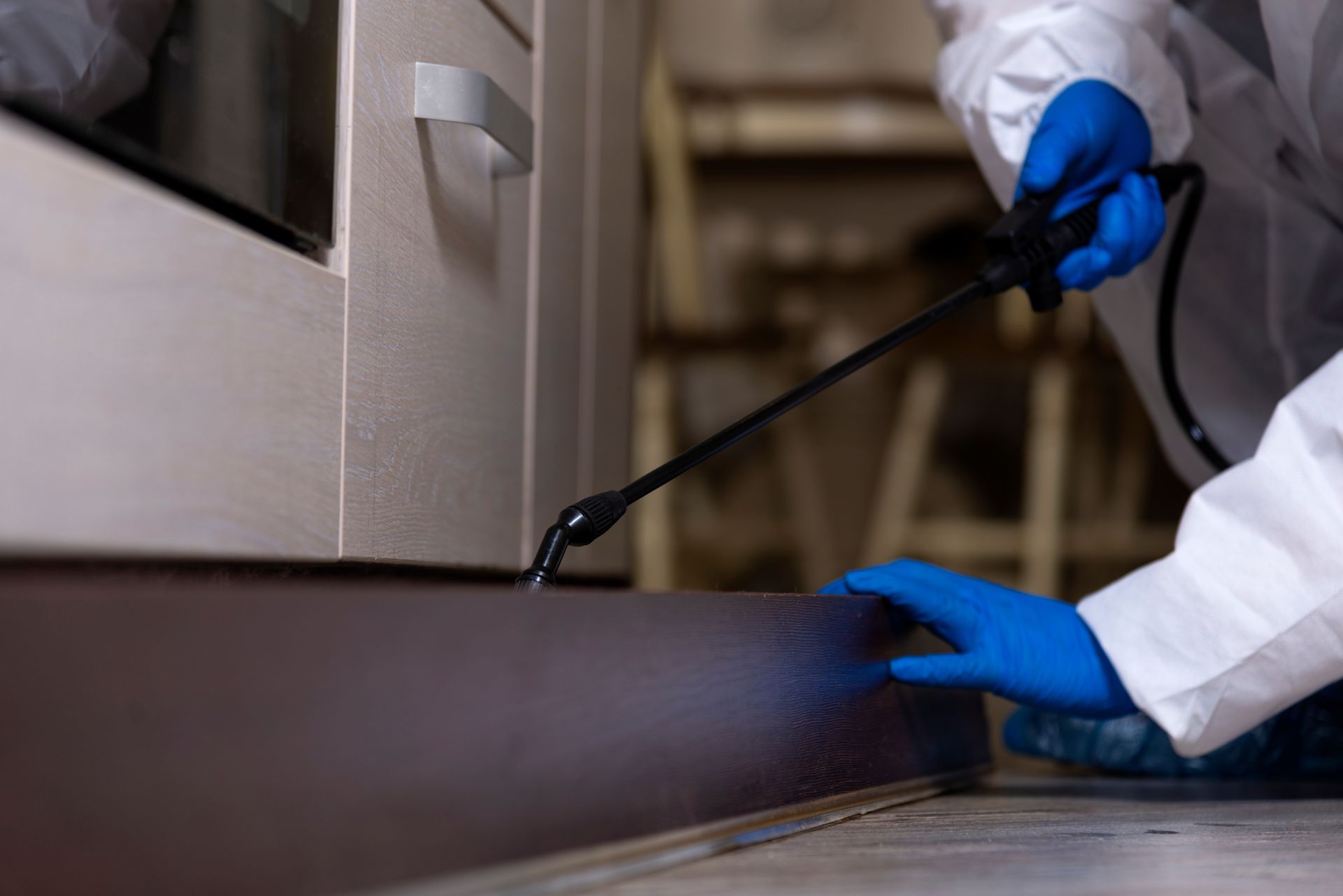 Person in protective suit spraying insecticide along a baseboard in a kitchen.