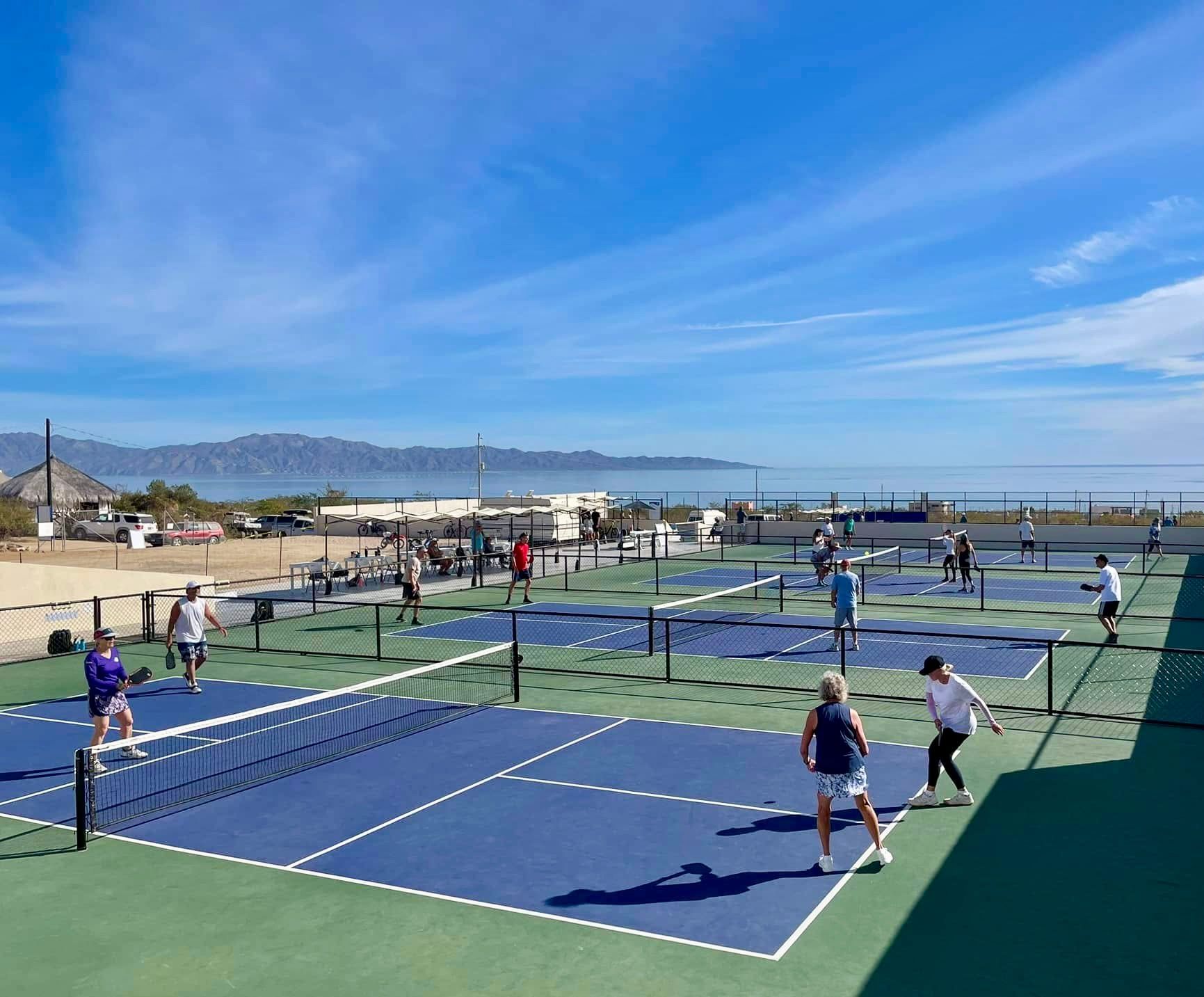 People playing pickleball on blue courts near the ocean, under a clear sky.