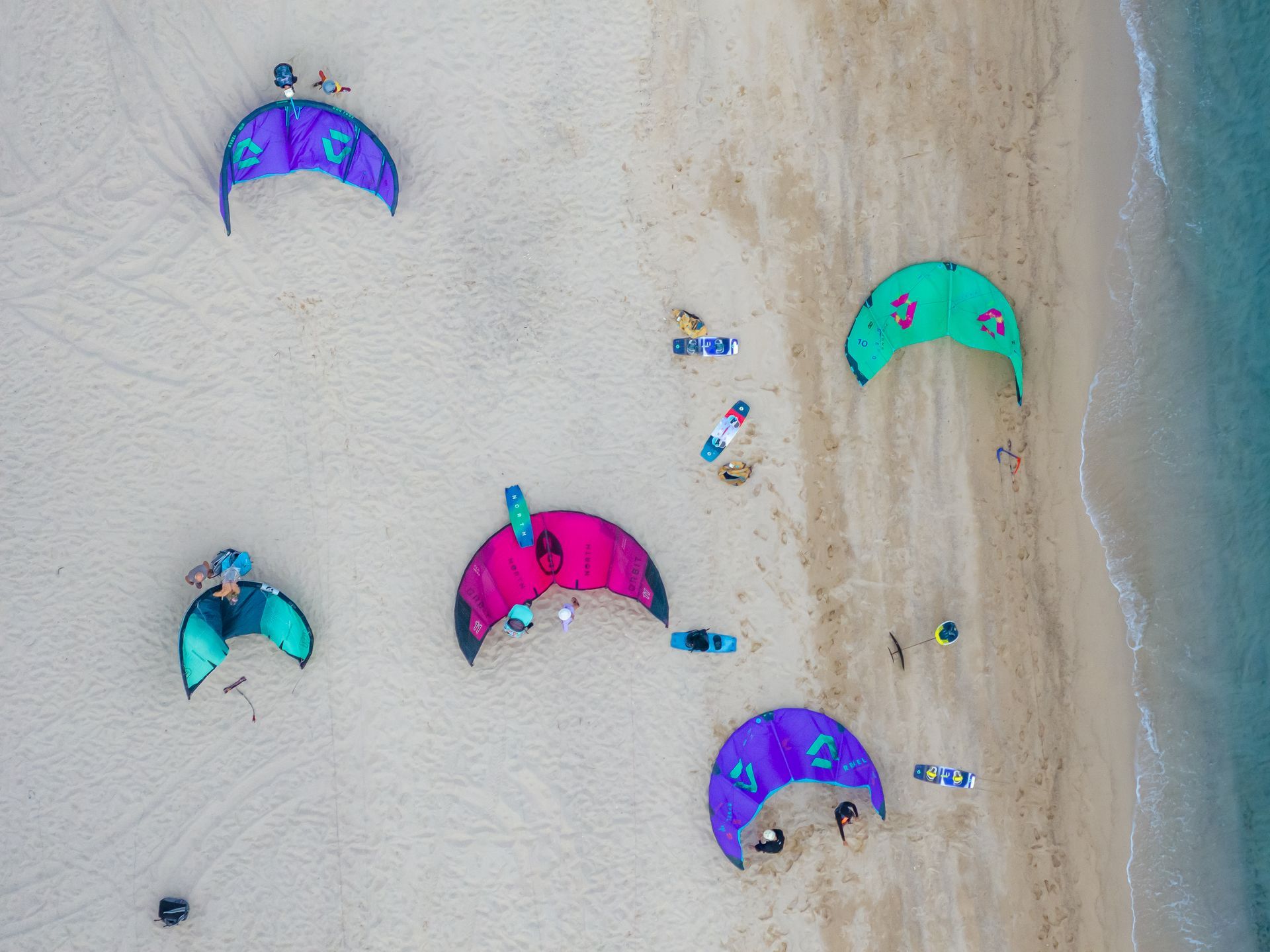 Aerial view of colorful wing foils on a sandy beach near the ocean. Several people are present.