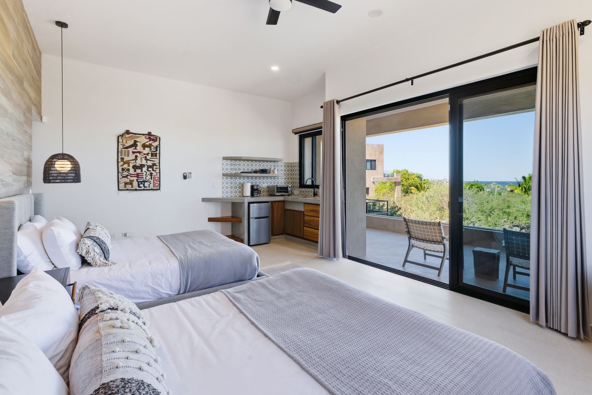 Two queen beds in a bright room with a small kitchenette and a sliding door leading to a balcony.