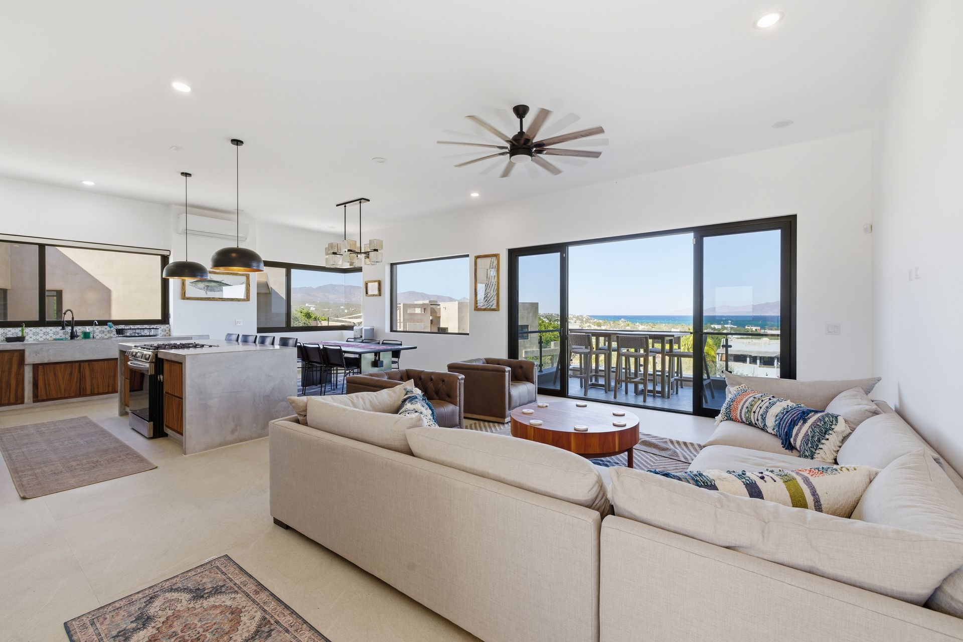 Open-plan living space with kitchen, dining, and seating areas; large windows with ocean view, neutral colors.