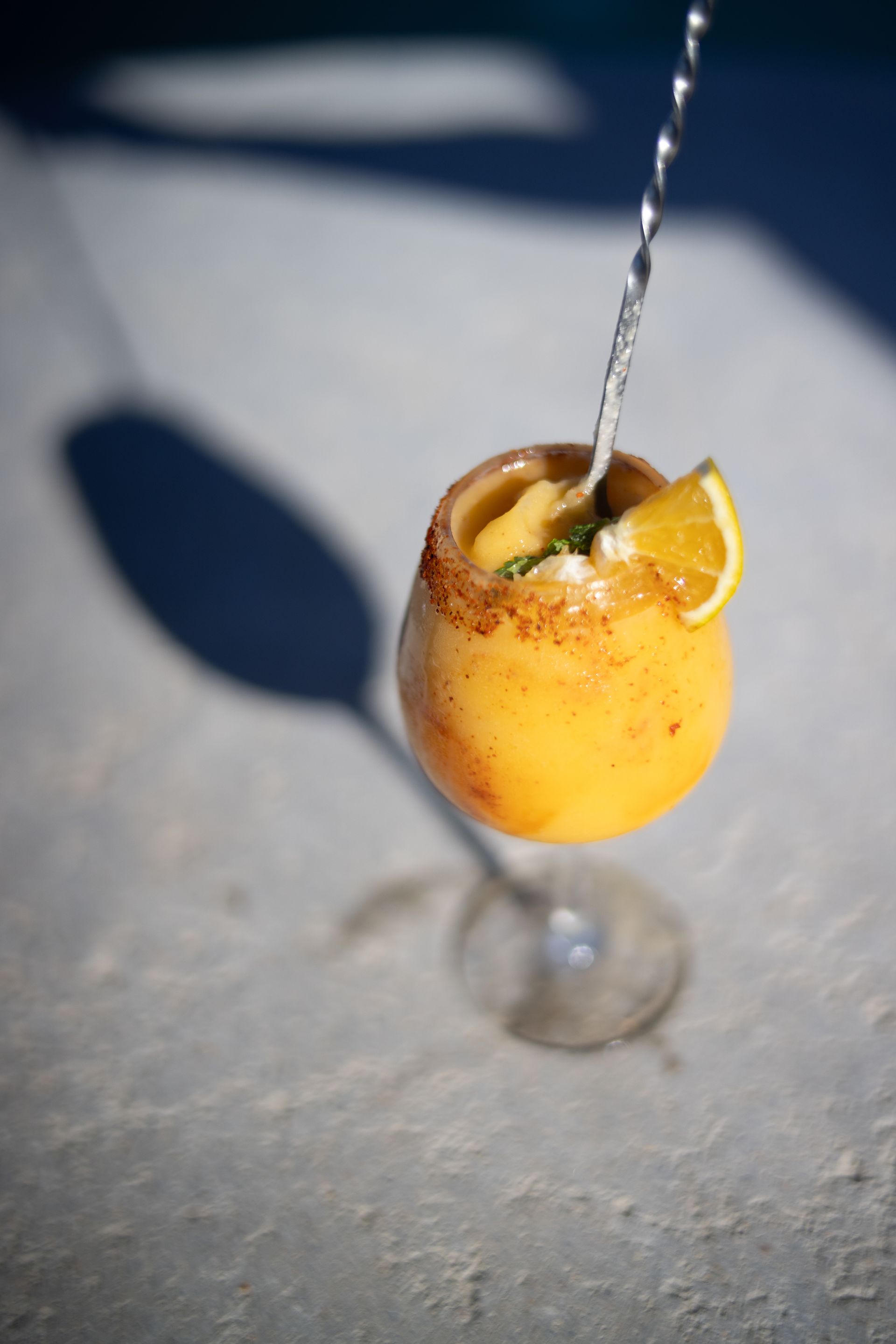 A yellow-orange cocktail in a stemmed glass with a citrus slice, being poured over ice.