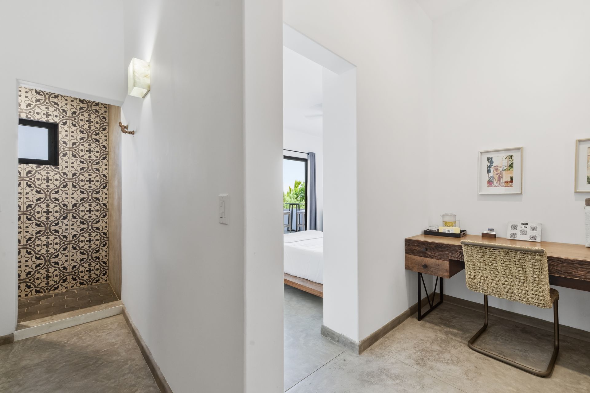 Hallway with shower, desk, and bedroom view. White walls, concrete floor, wooden desk, patterned shower tile.