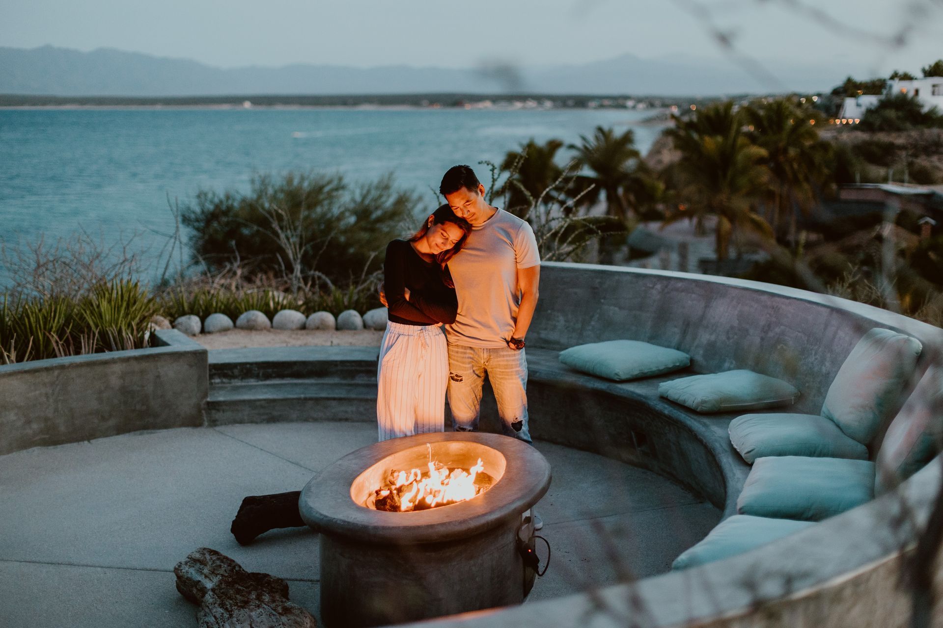 Couple embracing by a fire pit overlooking the ocean at dusk.