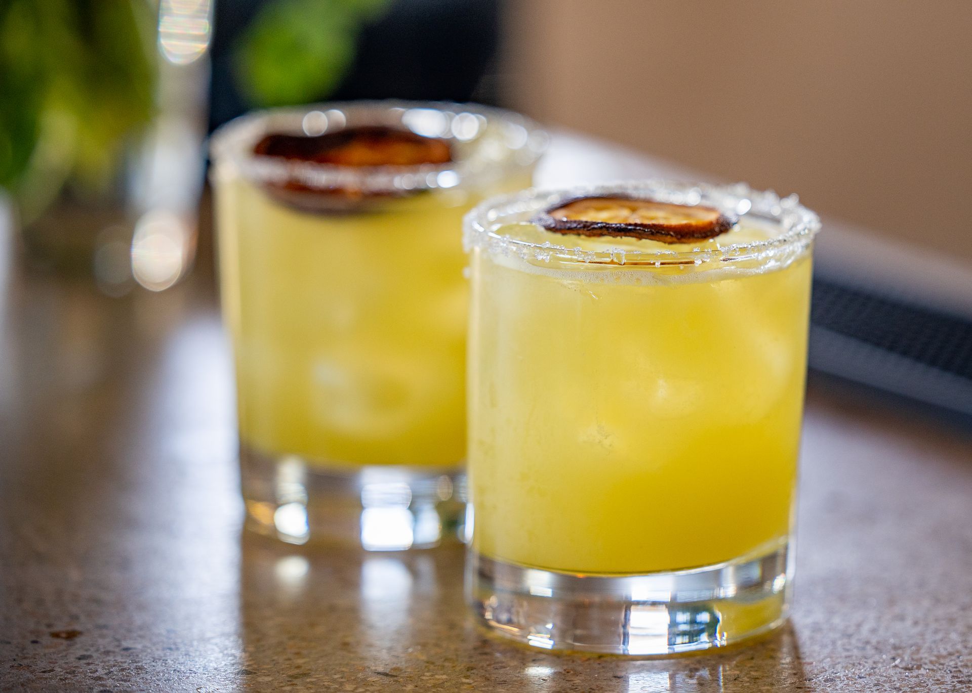 Two yellow cocktails in glasses with salt rims and a garnish, on a counter.
