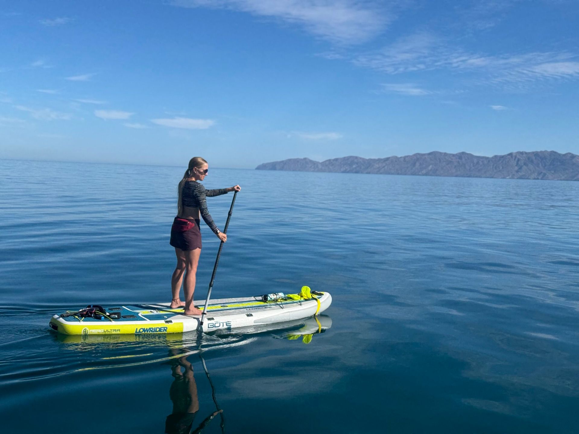 Woman paddles a stand-up board on calm ocean, wearing sun-protective clothing, with distant land in the background.