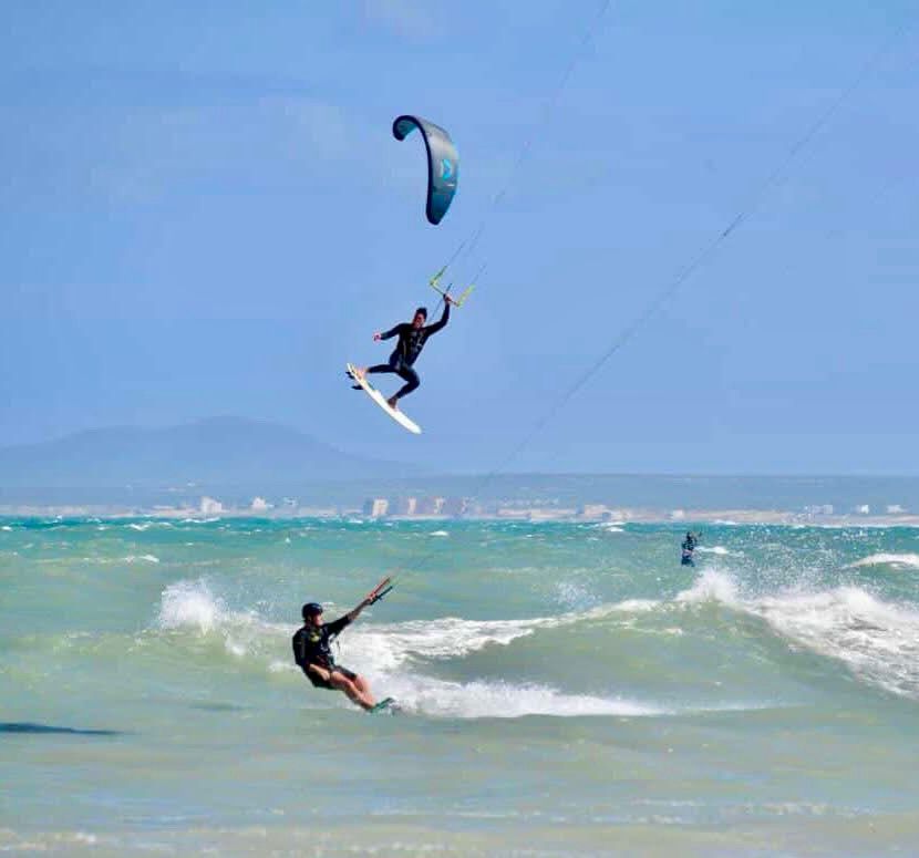 Kitesurfers on the water, one airborne. Blue kite and sky, white water, sunny day.