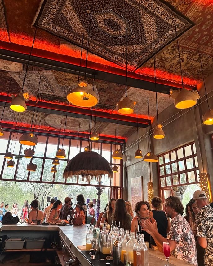 Bar with people; warm lighting, patterned ceiling, outdoor view.