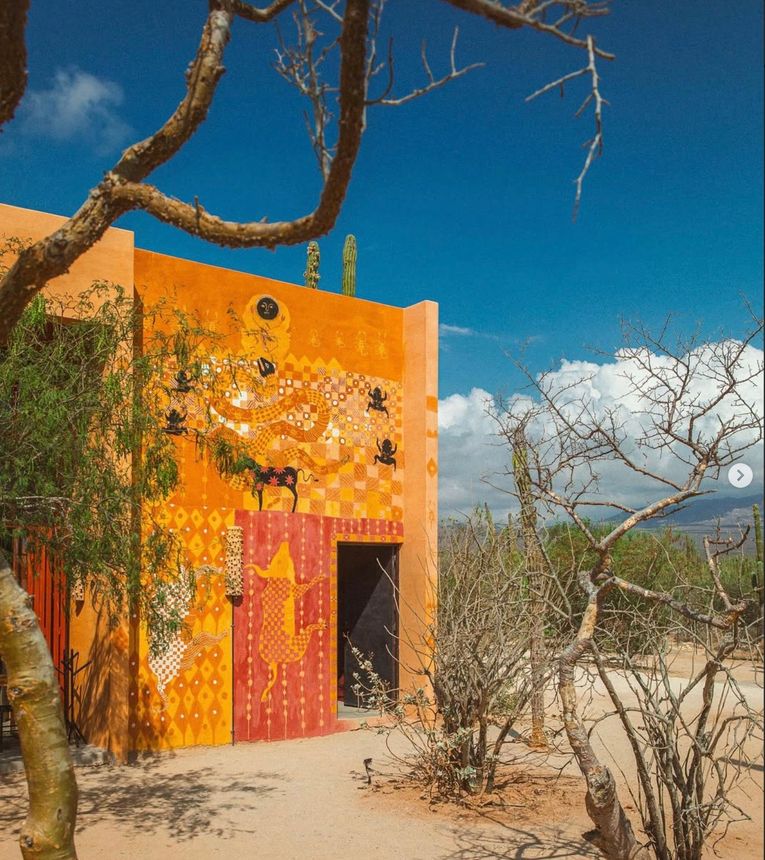Orange stucco building with open red door under a blue sky, framed by desert trees.