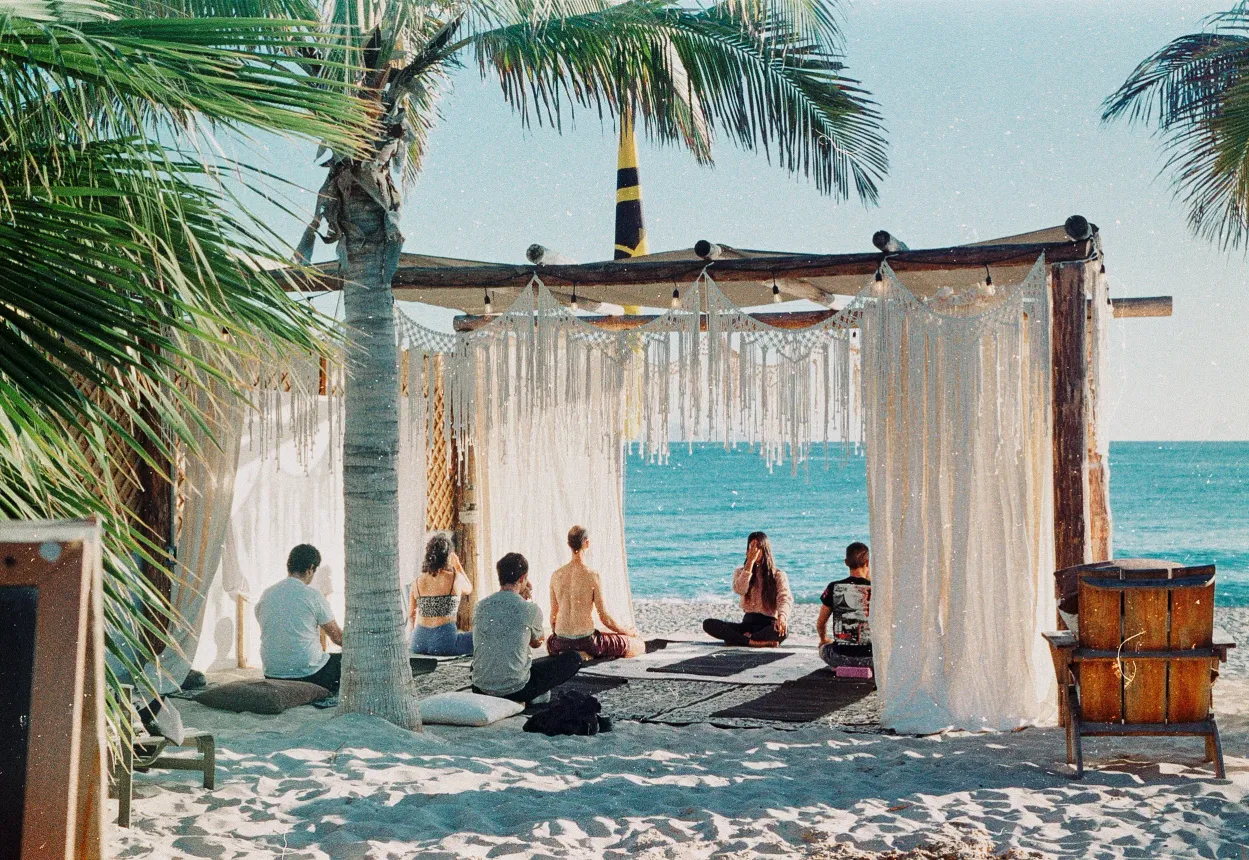 People meditating on a beach under a wooden canopy with white fringe, ocean in the background.