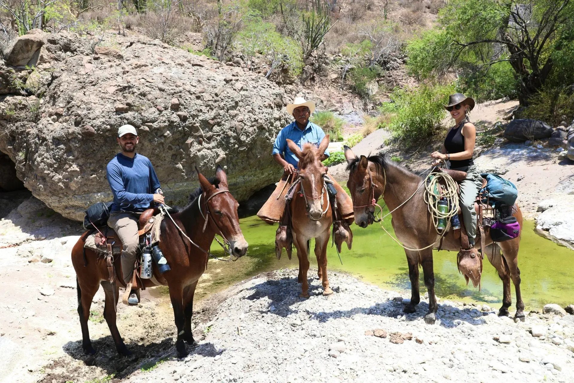 Three people on mules in a rocky, outdoor setting near a pool of water.
