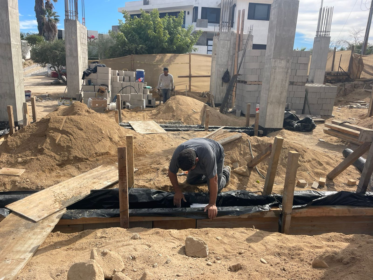 Construction site with worker kneeling over rebar in a foundation trench, surrounded by dirt and concrete posts.