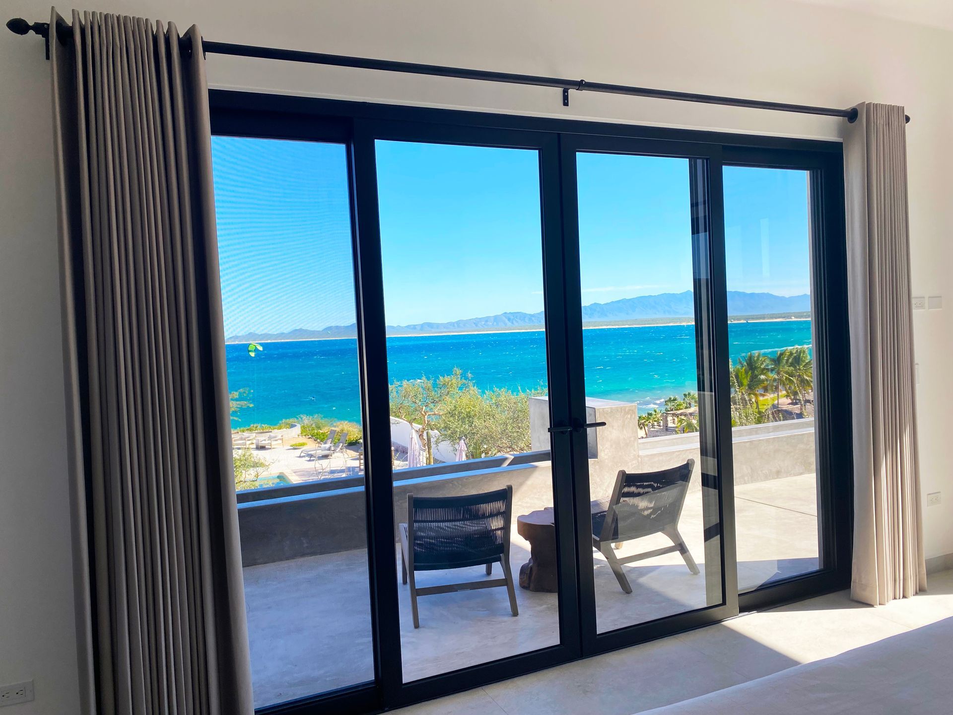 Sliding glass doors open to a beach view with seating on a patio. Blue ocean and sky.