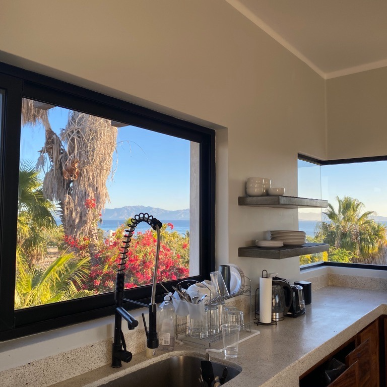 Kitchen with a large window overlooking a beach with palm trees and a view of the sea.