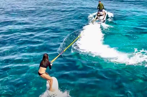 Woman on a hydrofoil being towed by a jet ski in blue water. Another person on a jet ski in the background.