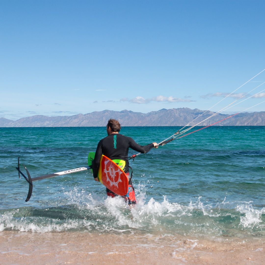 Person with kitesurfing equipment standing in shallow water, beach, blue sky.