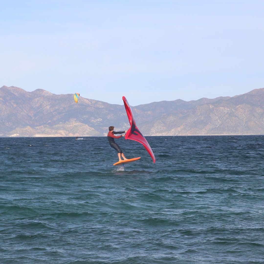 Person wing foiling on a board, red wing, blue water, mountains in the background.