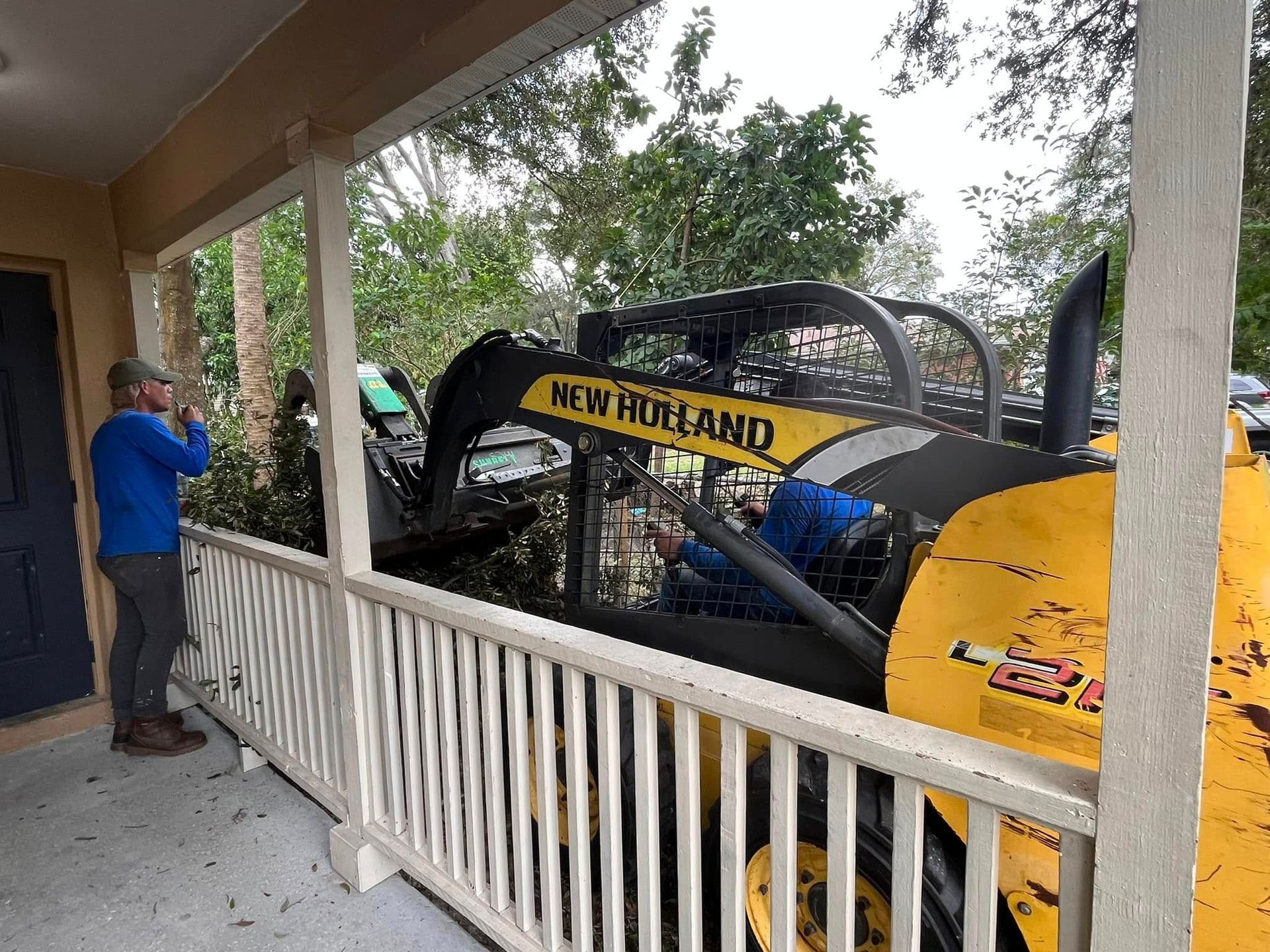 Man in blue shirt beside New Holland construction vehicle on a porch in a wooded area Man Construction Vehicle
