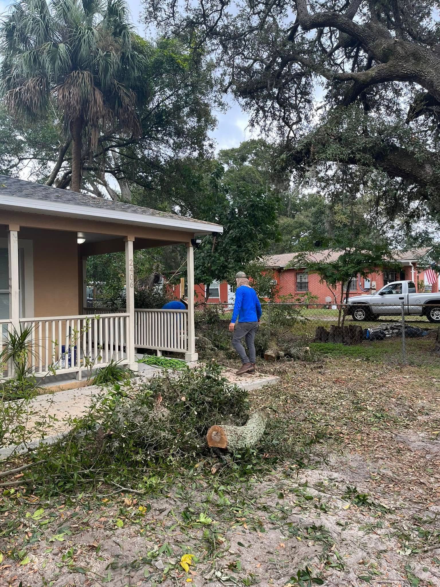 Yard with fallen tree branches, a person in a blue shirt, and a house with a porch after storm damage Land Clearing Services
