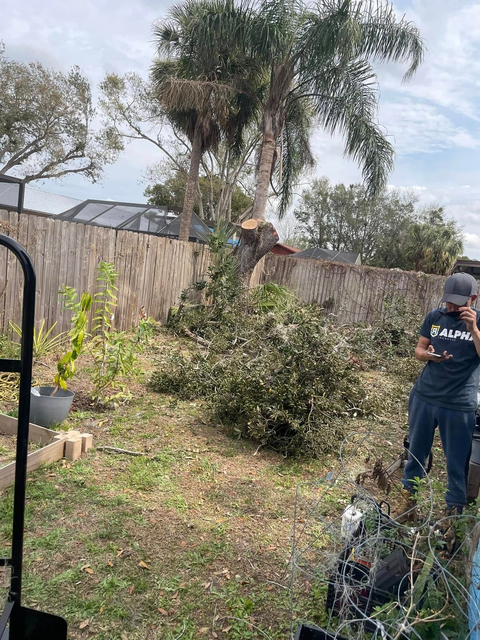 Person in navy work clothes trimming a shrub in a backyard beside a stone wall and trees. Tree Service Person