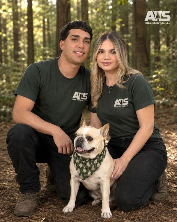 Two people in matching black shirts pose with a small dog in a forest setting. Owners