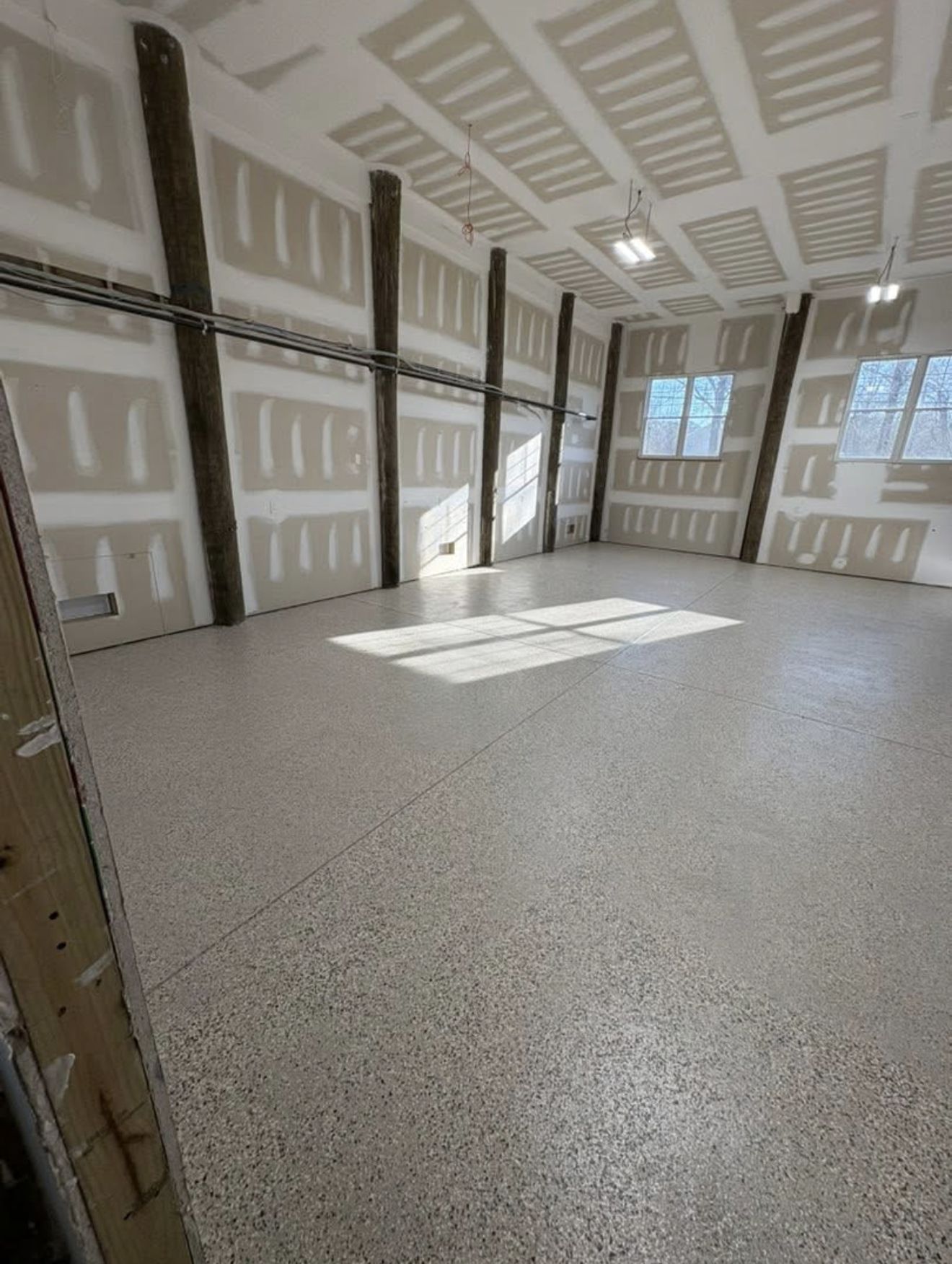 Empty interior with unfinished drywall walls and ceiling, concrete floor, wooden support beams, natural light.