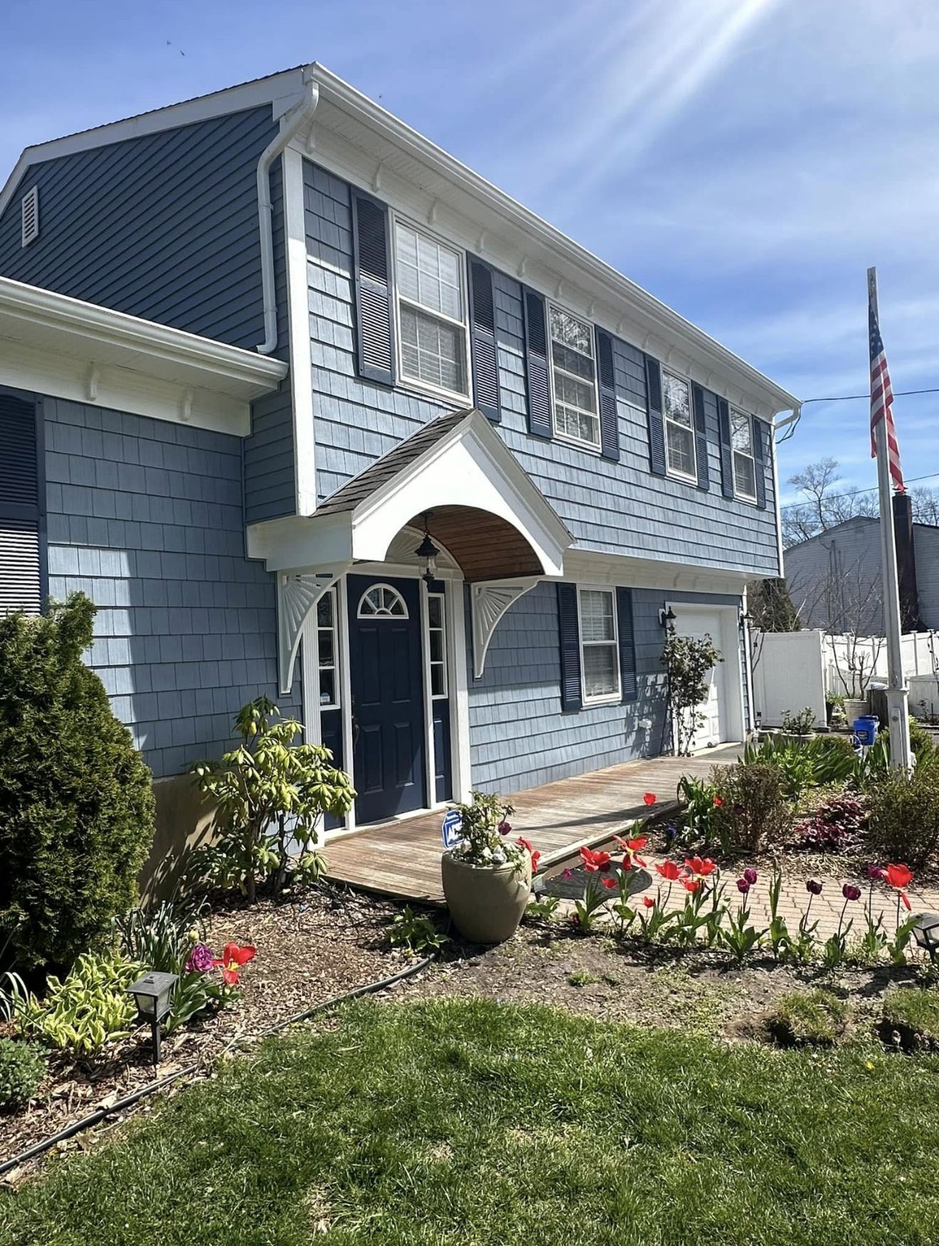 Blue two-story house with white trim, arched entryway, and American flag. Spring flowers in yard.