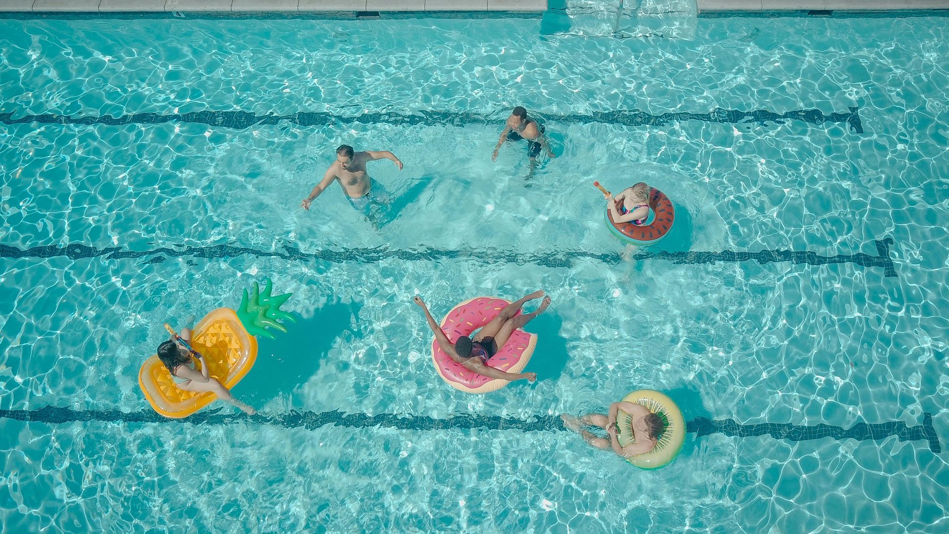 People floating in colorful inner tubes and swimming in a turquoise pool, viewed from above.