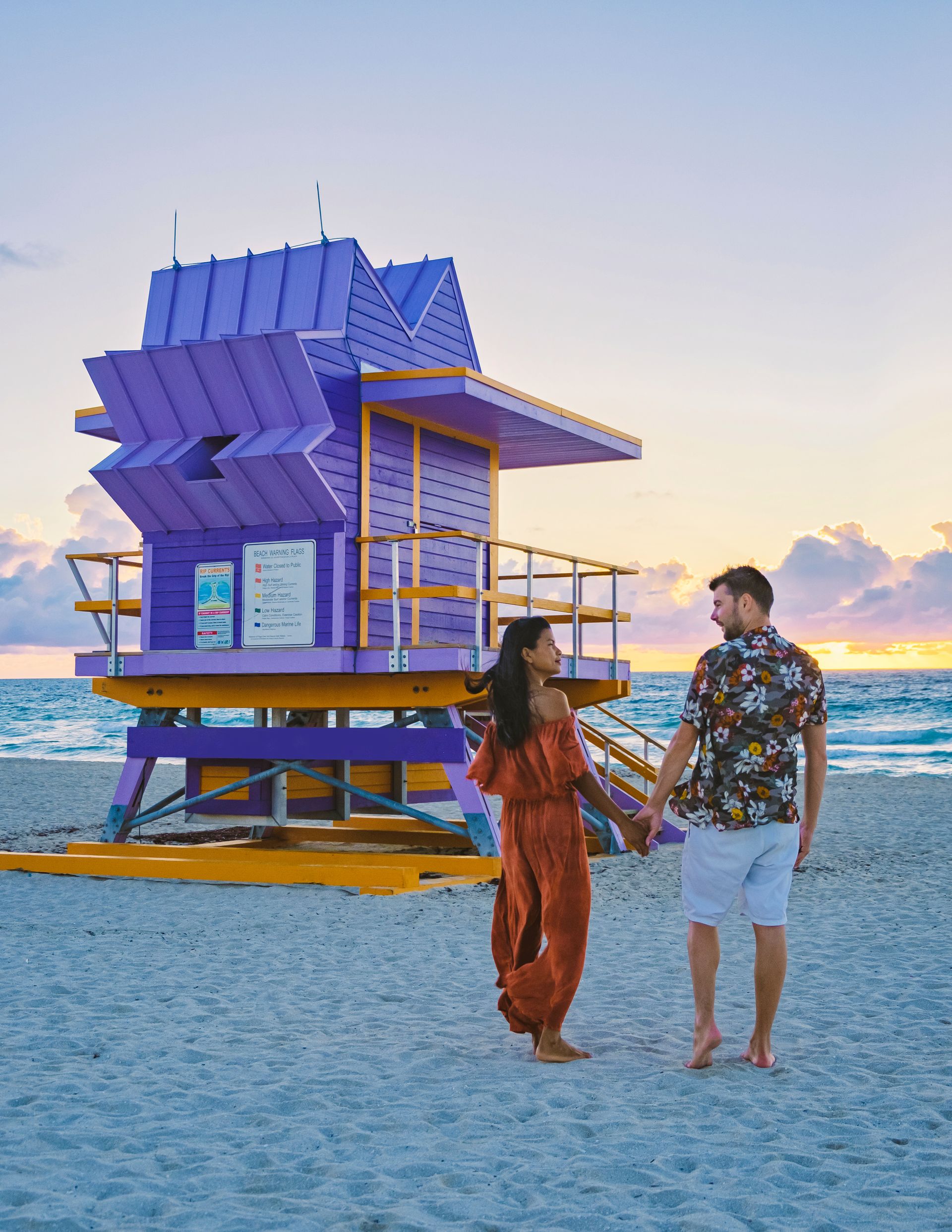 Couple holding hands on Miami Beach, walking toward a purple lifeguard stand at sunrise.
