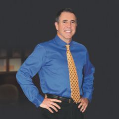 Man in blue shirt, yellow tie, hands on hips, smiling, against a dark background.