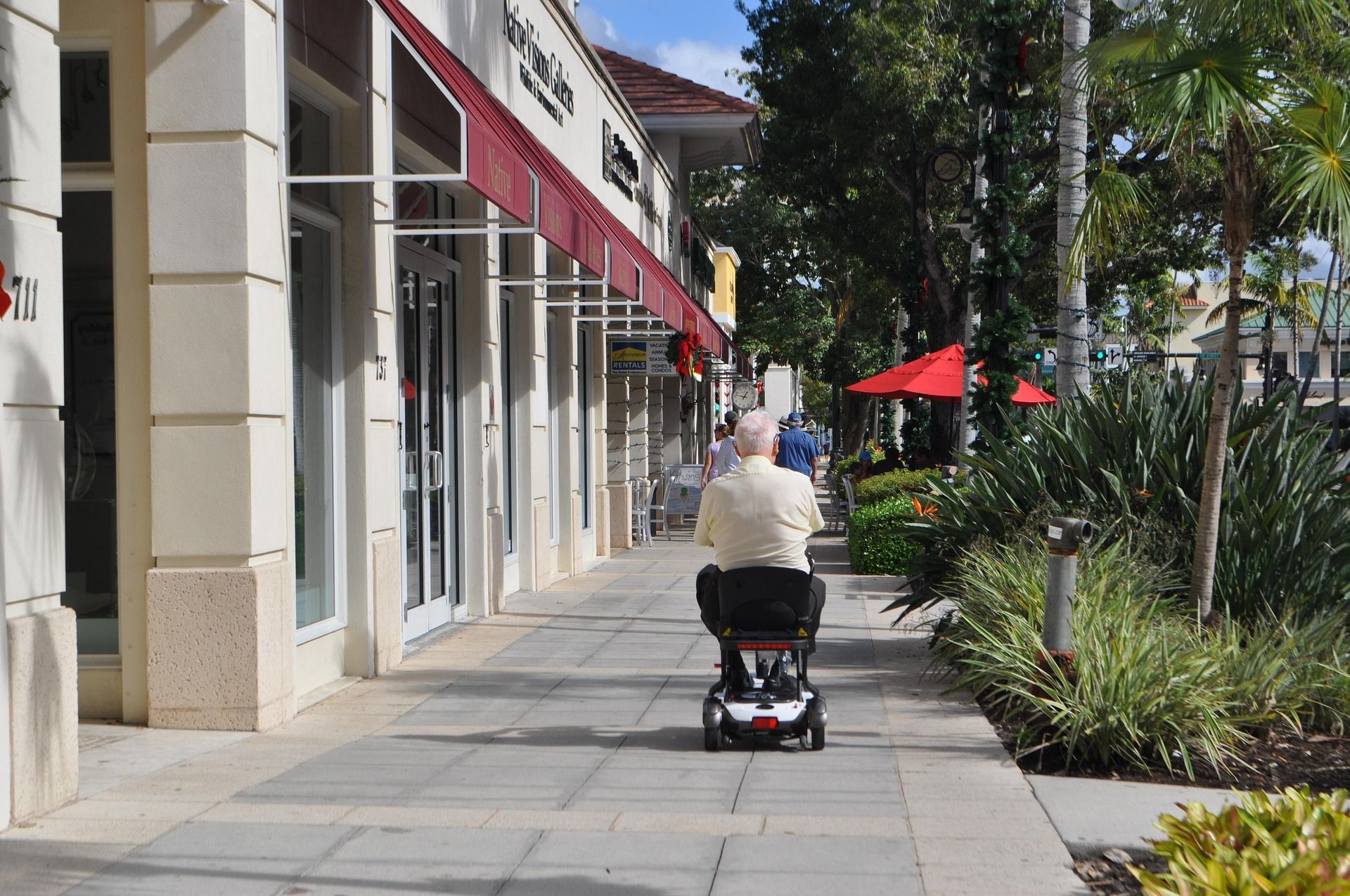 Person on motorized scooter on a sidewalk lined with shops under awnings, palm trees, and sunny sky.