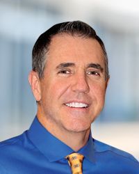 Man in blue shirt and patterned tie smiling; blurred background.