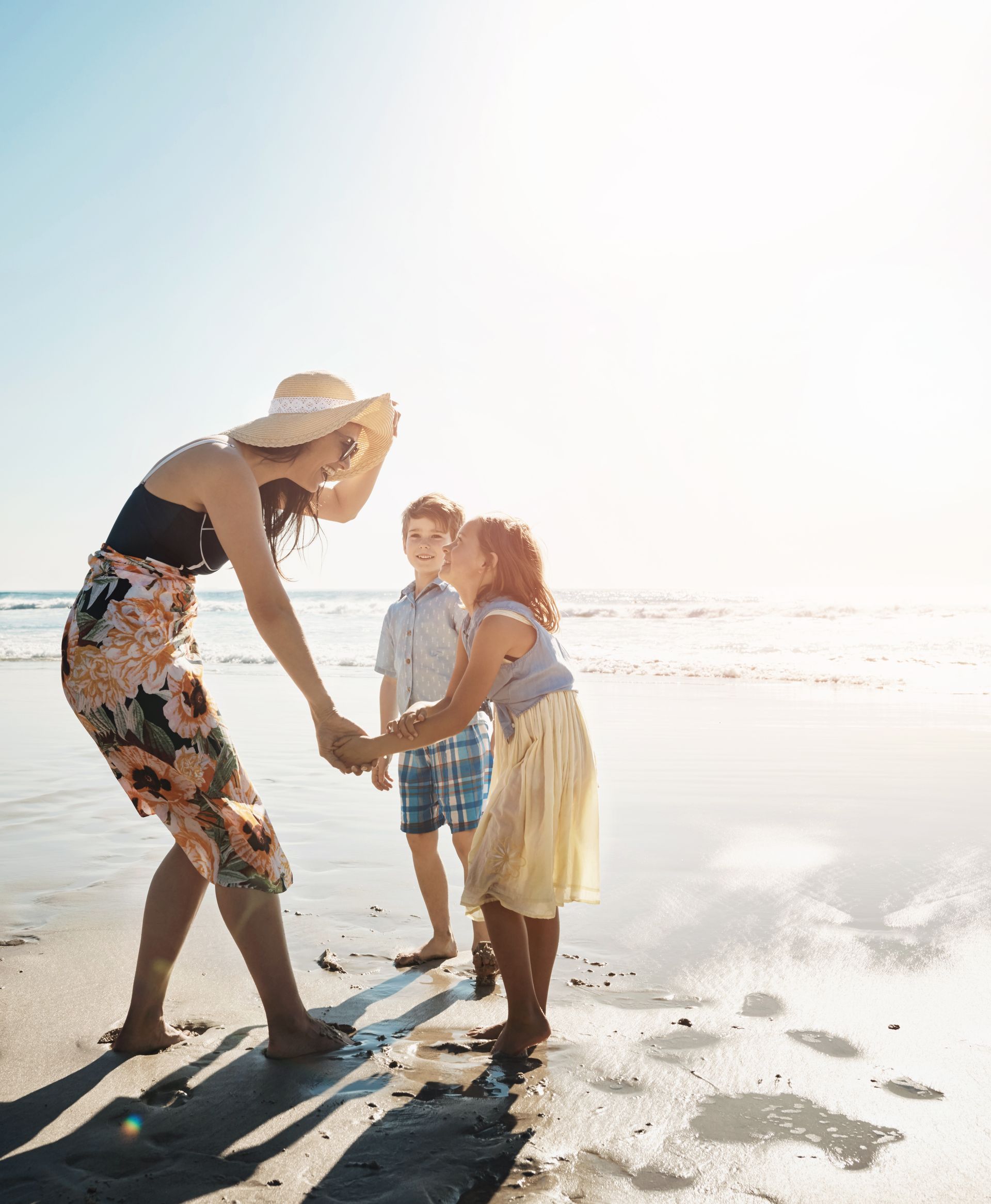 Woman and two children play on a beach, holding hands. Bright sunlight, water in the background.