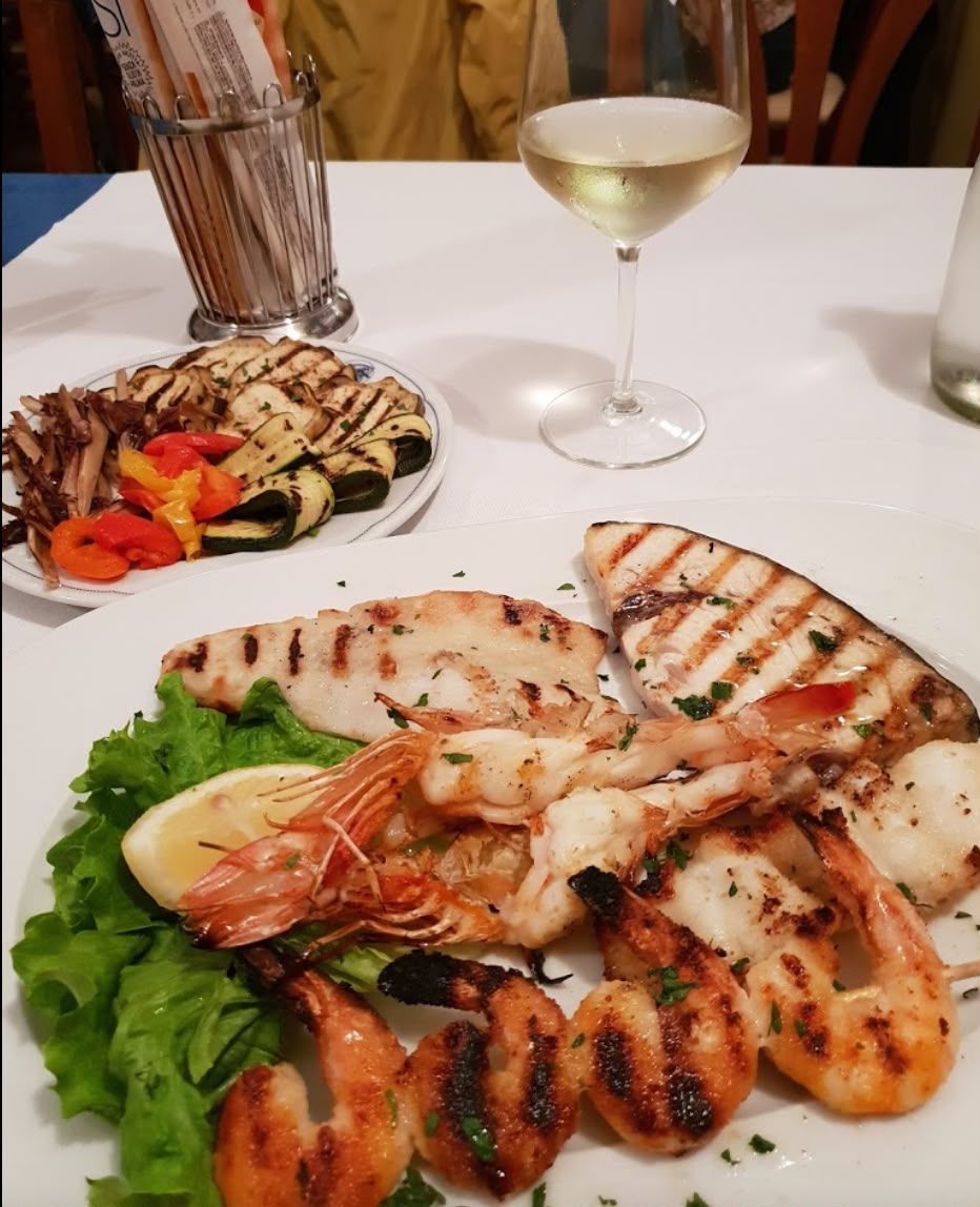 Piatto di frutti di mare alla griglia con gamberi e capesante, accompagnato da verdure e un calice di vino bianco.