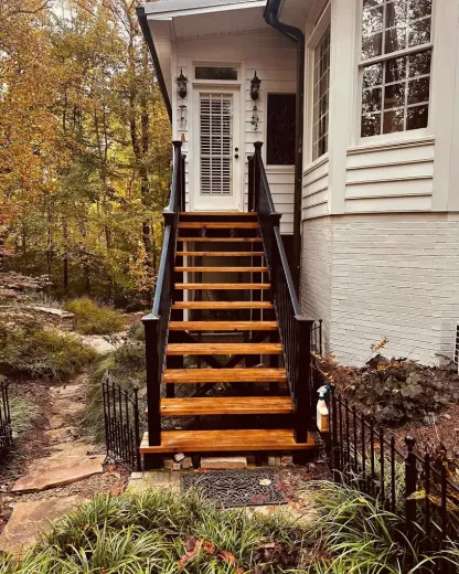 Wooden front stairs leading to a white house entrance, bordered by black railings and autumn foliage.