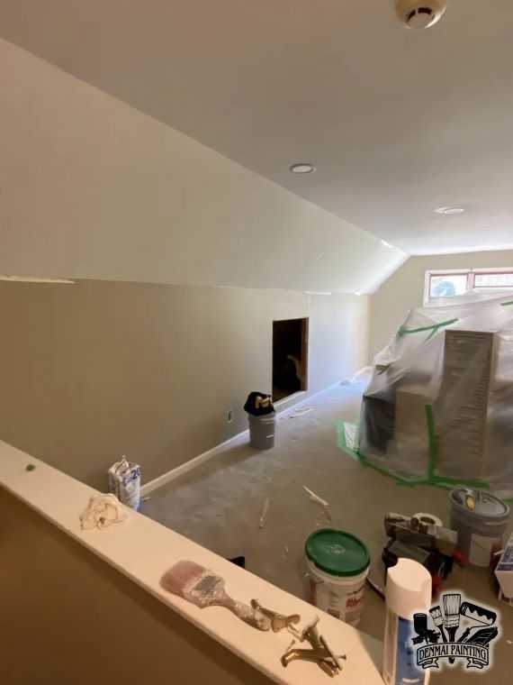 Interior under renovation with taped furniture, paint supplies, and a sloped ceiling in a small room