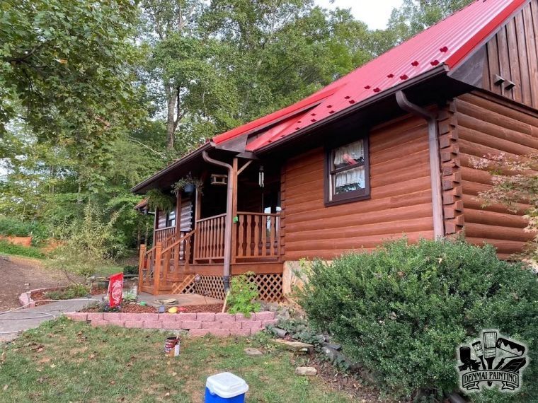 Log cabin with red roof and porch, surrounded by trees and shrubs in a wooded yard.
