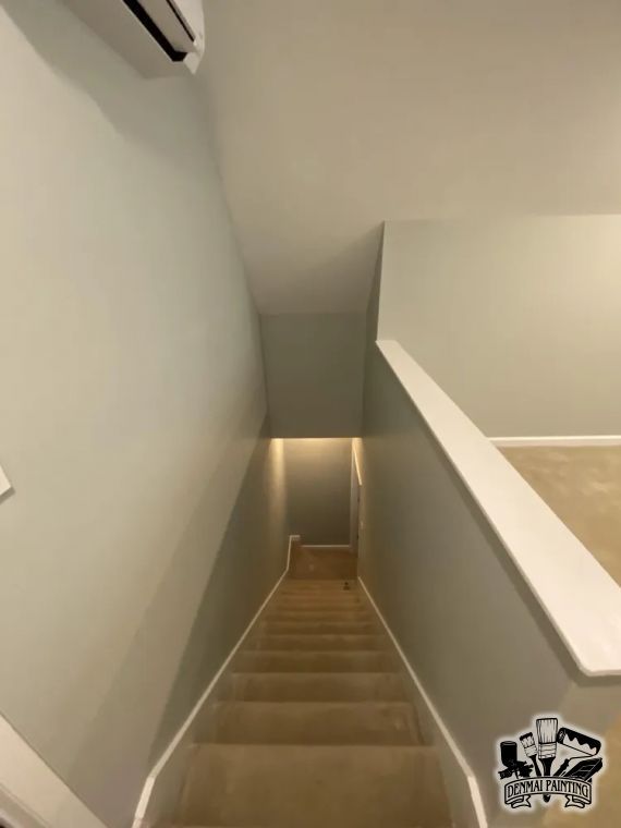 Narrow stairwell with beige walls and a lit landing at the bottom, viewed from the top of the stairs