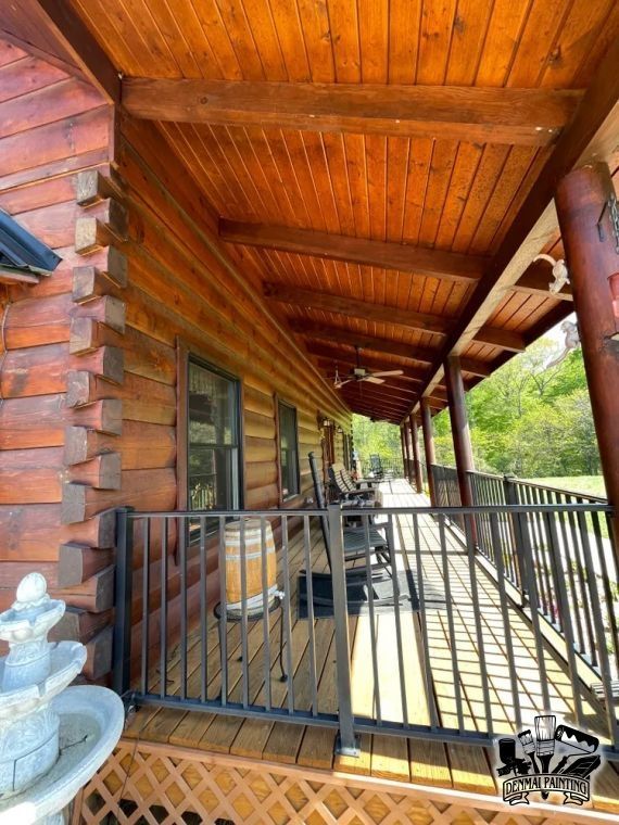Covered wooden porch with log walls, black railing, and a sloped timber ceiling overlooking trees